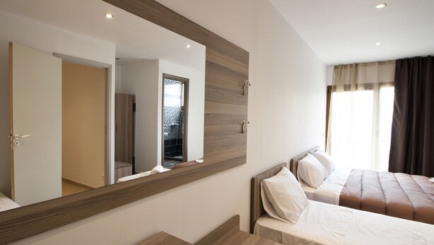 A modern hotel room with a large mirror, wooden furniture, a bed with white linens, and a view of the outside through a window with curtains. - Pension Pineapple (Skiathos)