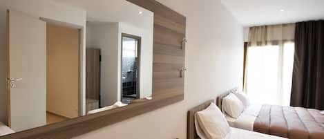 A modern hotel room with a large mirror, wooden furniture, a bed with white linens, and a view of the outside through a window with curtains.
