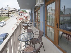 A balcony with wicker chairs and a glass table, overlooking a street with parked cars and buildings. - Pension Pineapple (Skiathos)