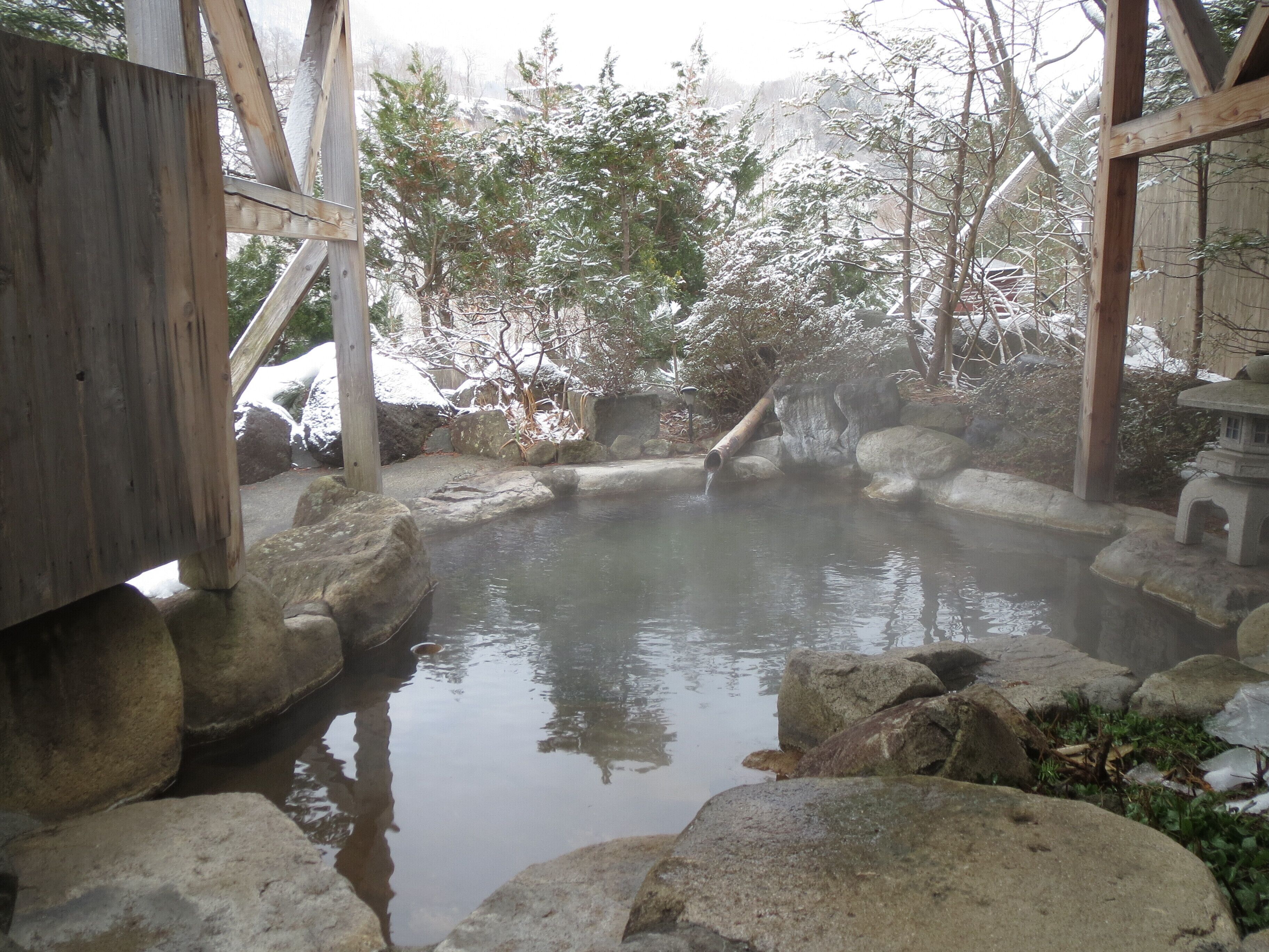 A traditional Japanese onsen with a wooden structure, a stone bath, and a natural setting with trees and rocks.