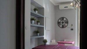 Un comedor con una mesa dispuesta para cuatro, sillas blancas, un reloj en la pared y estantes con plantas en macetas.