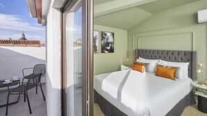A hotel room with a large bed, two chairs, a small table, and a view of the cityscape. - L&H La Latina (Madrid)