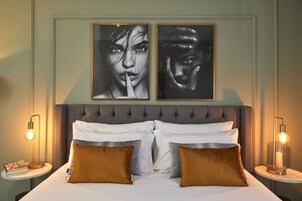 Ein ordentlich bezogenes Bett mit weißen Leinentüchern und zwei braunen Kissen, das von zwei Nachttischen mit Lampen flankiert wird, über dem Bett hängen zwei gerahmte Kunstwerke.