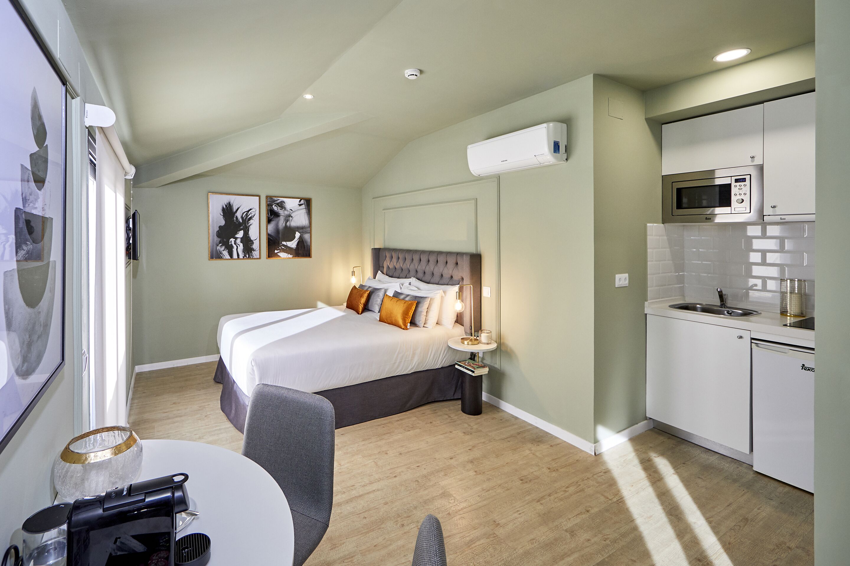 A modern hotel room with a bed, bedside table, and a kitchenette.