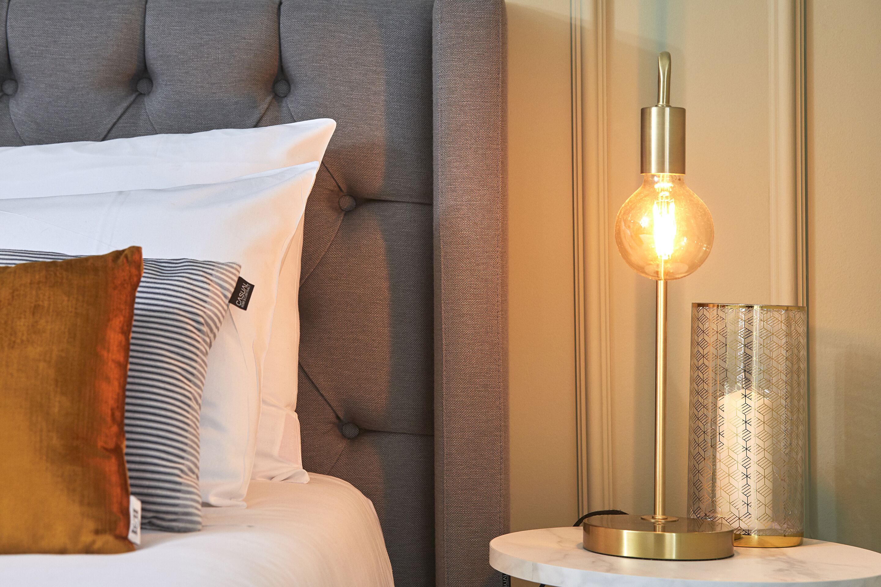 A neatly made bed with a tufted headboard, a bedside table with a lamp, and a decorative glass container.