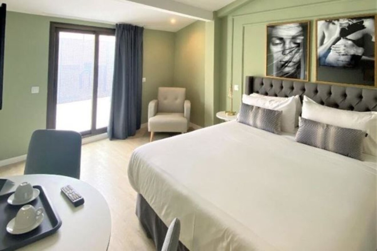 A hotel room with a large bed, a chair, a desk with a tray, and a large window with curtains.