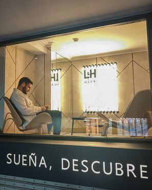 A man sitting in a chair at a table with a laptop, in a room with a large window and a sign that reads "SUEÑA, DESCUBRE". - L&H La Latina (Madrid)