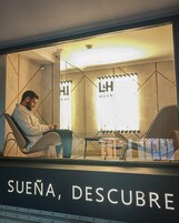 Ein Mann sitzt an einem Tisch mit einem Laptop, in einem Zimmer mit einem großen Fenster und einem Schild mit der Aufschrift 'SUEÑA, DESCUBRE'.