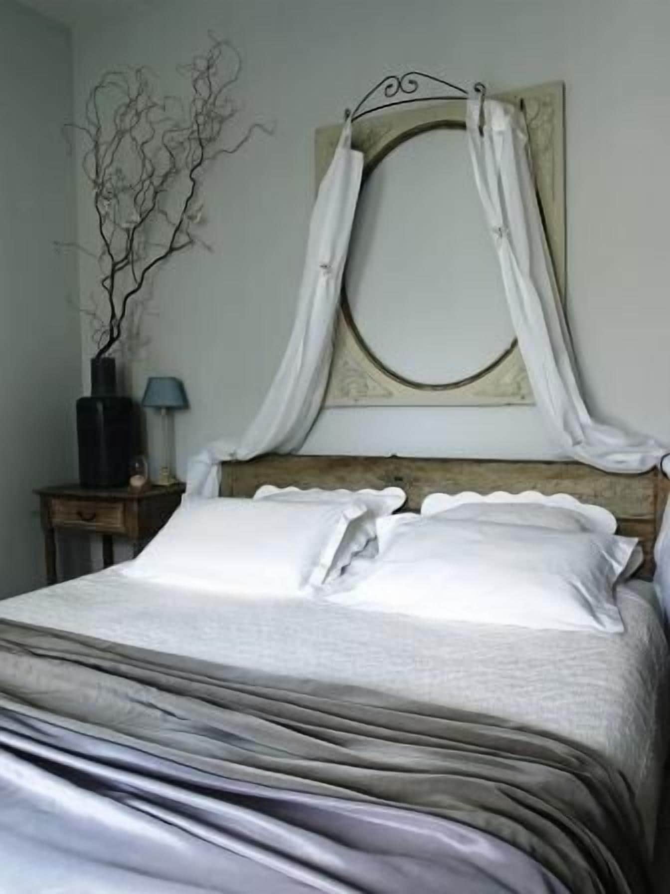 A neatly made bed with white bedding, a bedside table with a lamp, and a wall-mounted mirror with a decorative frame.