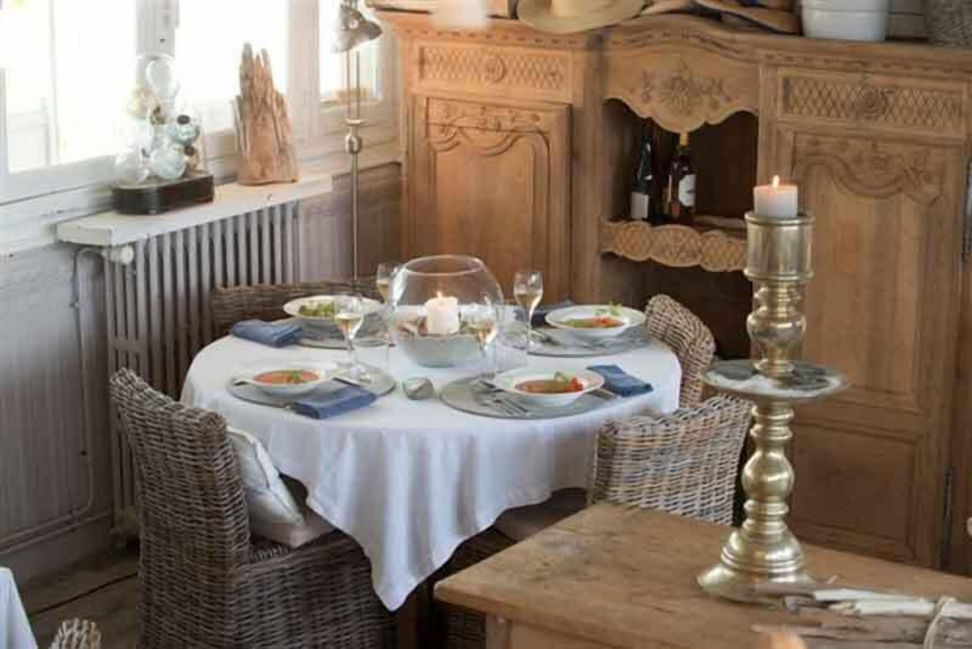 A dining area with a round table set for four, surrounded by wicker chairs. A white tablecloth covers the table, and there are plates, glasses, and a candle holder on it.