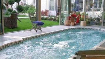 En indendørs swimmingpool med et boblebad, omgivet af en terrasse med siddepladser og udsigt til en have samt en bygning i baggrunden.