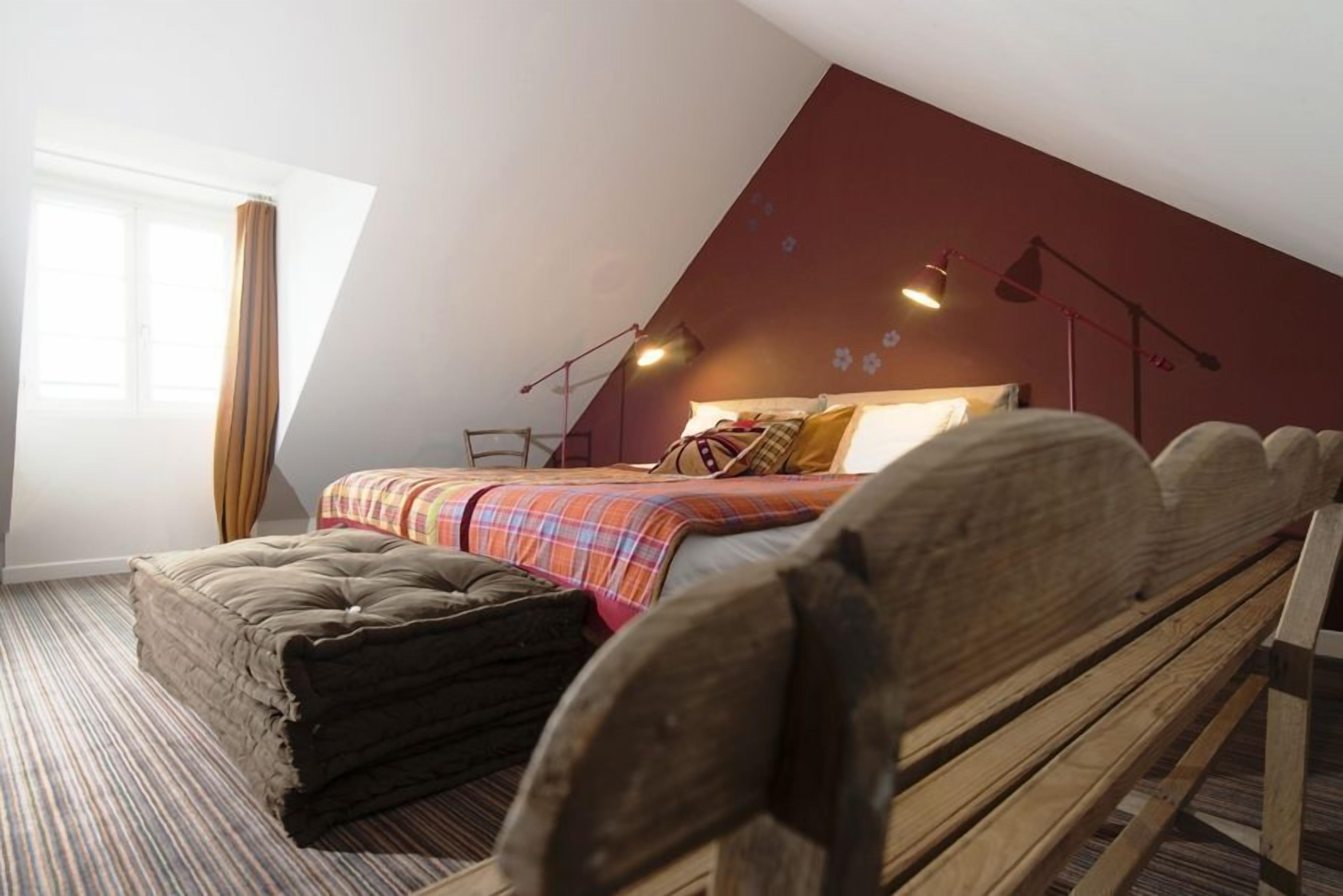A bedroom with a sloped ceiling, a bed with a plaid bedspread, a wooden bench, and a window with curtains.