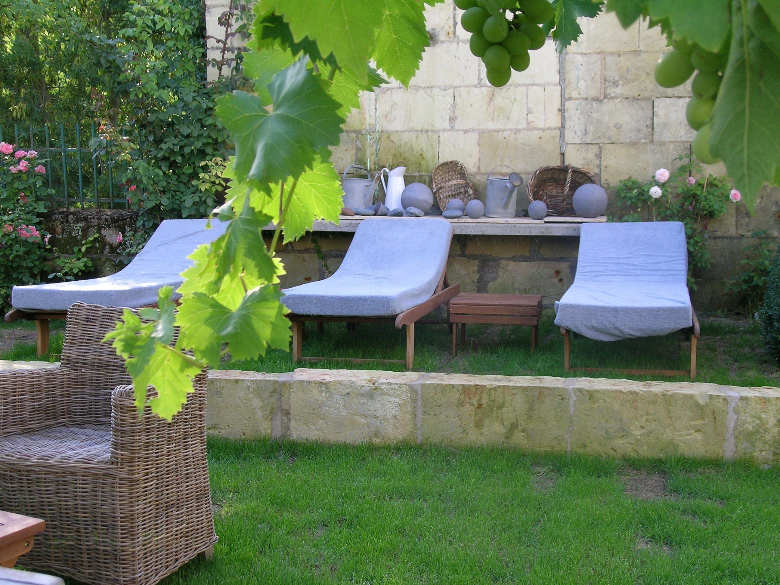 Un jardin agrémenté de trois chaises longues et d’un fauteuil en osier, d’un mur en pierre, ainsi que de diverses plantes et fleurs.