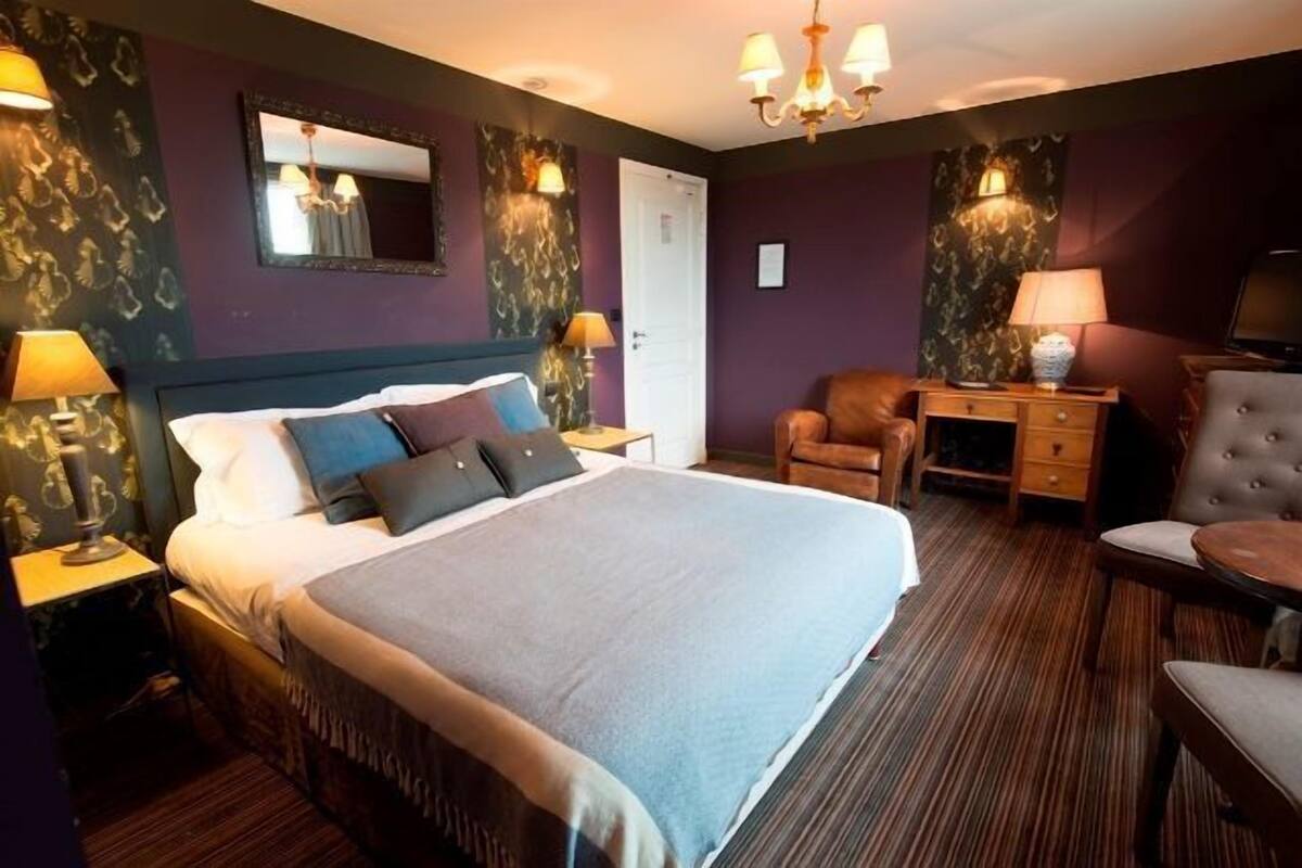 Habitació d'hotel amb un llit gran, dues taules de nit amb làmpades, una cadira, una taula petita i un mirall a la paret.
