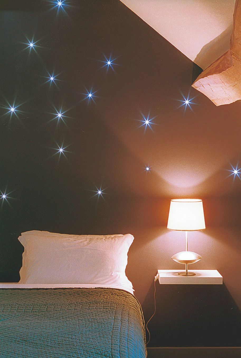 Un dormitorio con una cama, una mesita de noche con una lámpara y adhesivos decorativos con forma de estrella en la pared.