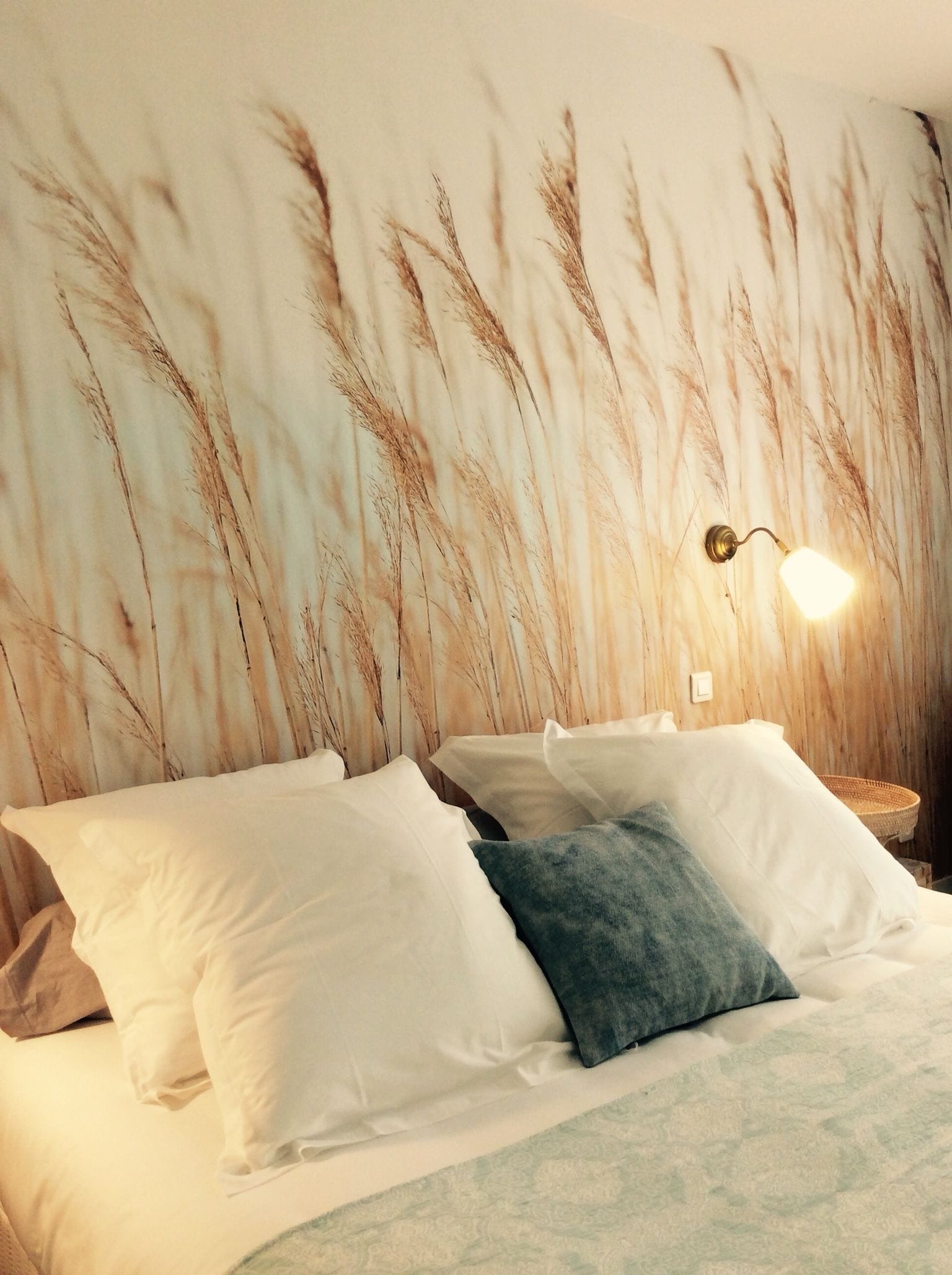 Una cama con almohadas blancas y azules, una pared con un mural de un campo de trigo y una lámpara de pared.