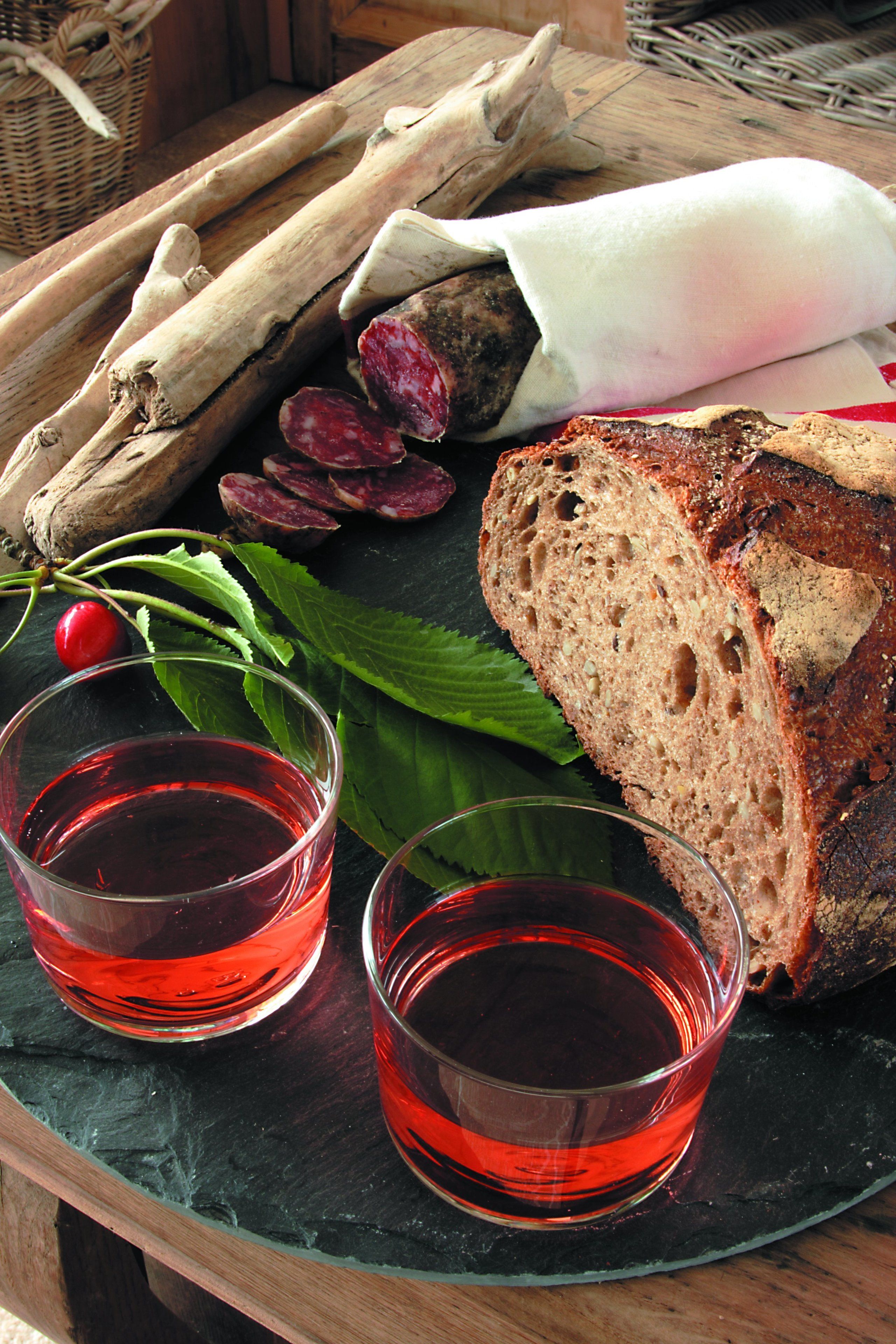 A wooden tray with sliced bread, a whole baguette, fresh herbs, and two glasses of red liquid.