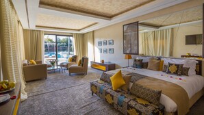 Ein geräumiges Hotelzimmer mit einem großen Bett, einem Sofa, einem Sessel und einem kleinen Tisch. Es gibt ein großes Fenster mit Vorhängen, einen Flachbildfernseher und einen wandmontierten Spiegel.