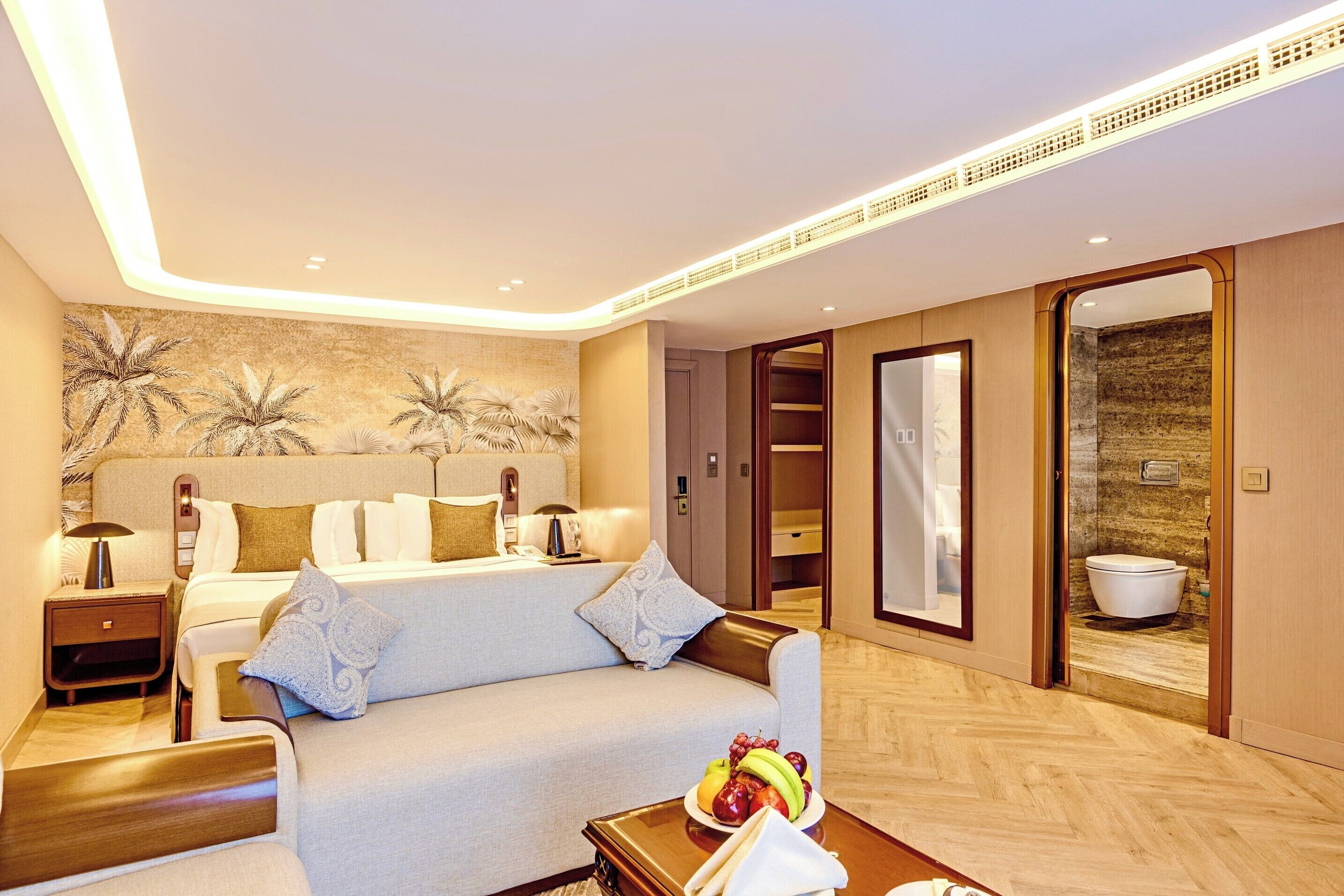 Une chambre d’hôtel avec un lit, un canapé, une table de chevet avec une lampe, un panier de fruits et une salle de bain visible à travers une porte ouverte.