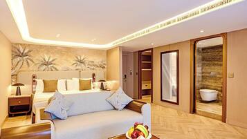 Une chambre d’hôtel avec un lit, un canapé, une table de chevet avec une lampe, un panier de fruits et une salle de bain visible à travers une porte ouverte.