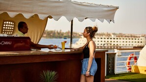 A bar area with a wooden counter, a person serving a drink, and a view of a body of water and buildings in the background. - Sonesta St George Nile Cruise  Ship - 3 & 7 Nights - From  Aswan  Every Friday (Luxor)