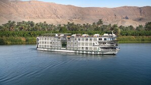 A large river cruise ship with multiple decks and balconies, floating on a calm river with palm trees and mountains in the background. - Sonesta St George Nile Cruise  Ship - 3 & 7 Nights - From  Aswan  Every Friday (Luxor)