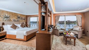 A modern hotel room with a large bed, a sitting area with a view of the water, and a balcony. - Sonesta St George Nile Cruise  Ship - 3 & 7 Nights - From  Aswan  Every Friday (Luxor)