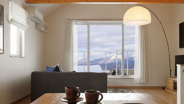 Ein Zimmer mit Esstisch, zwei Tassen, einem Schachbrett, einer Pflanze, einem Sofa, einem Fenster mit Bergblick und einer Pendelleuchte.