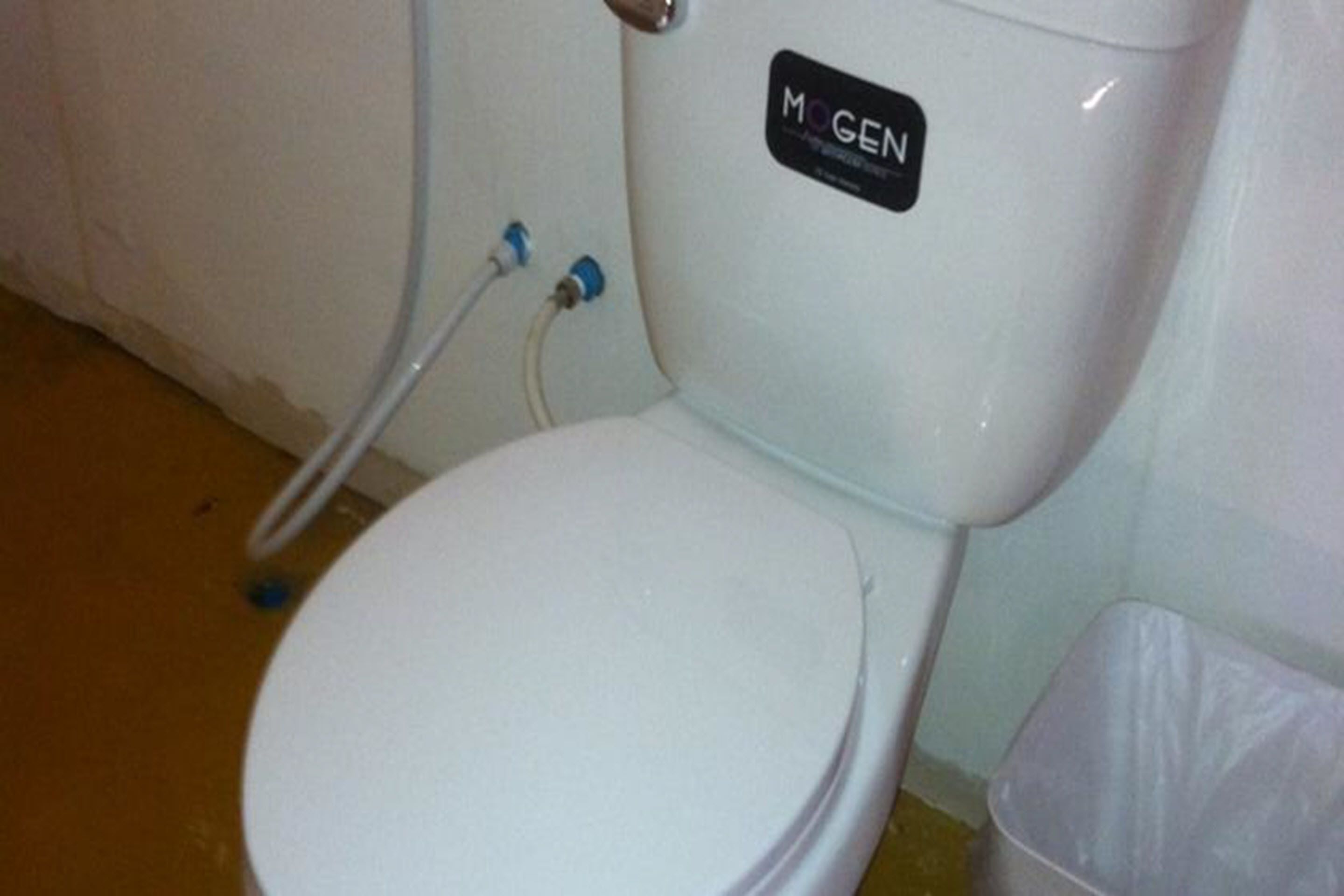 A white toilet with a "MOGEN" label, a plastic trash bin, and a white wall.
