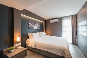 A modern hotel room with a large bed, a bedside table with a lamp, a wall-mounted air conditioner, and a window with curtains. - Fyn Hotel (Bangkok)