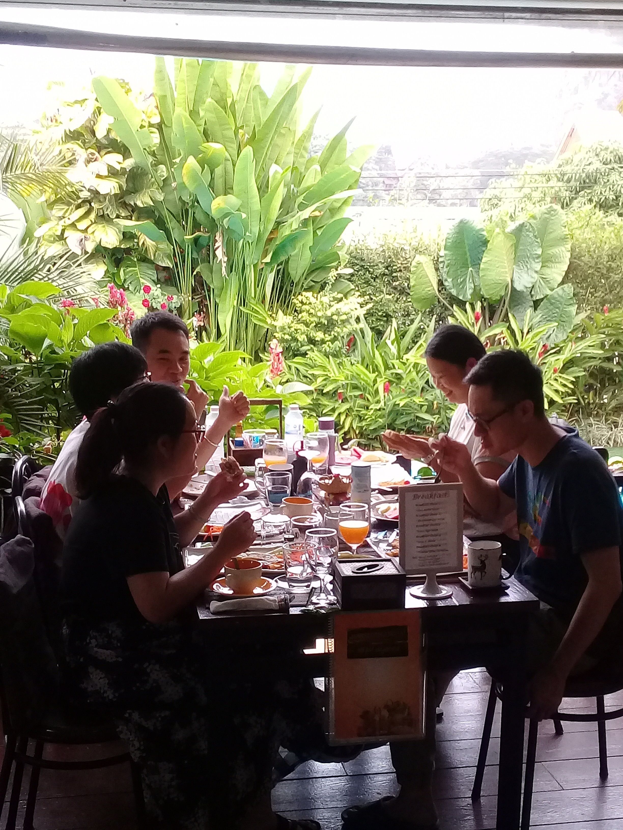 Um grupo de pessoas jantando em uma mesa ao ar livre com um cardápio, cercado por vegetação exuberante.