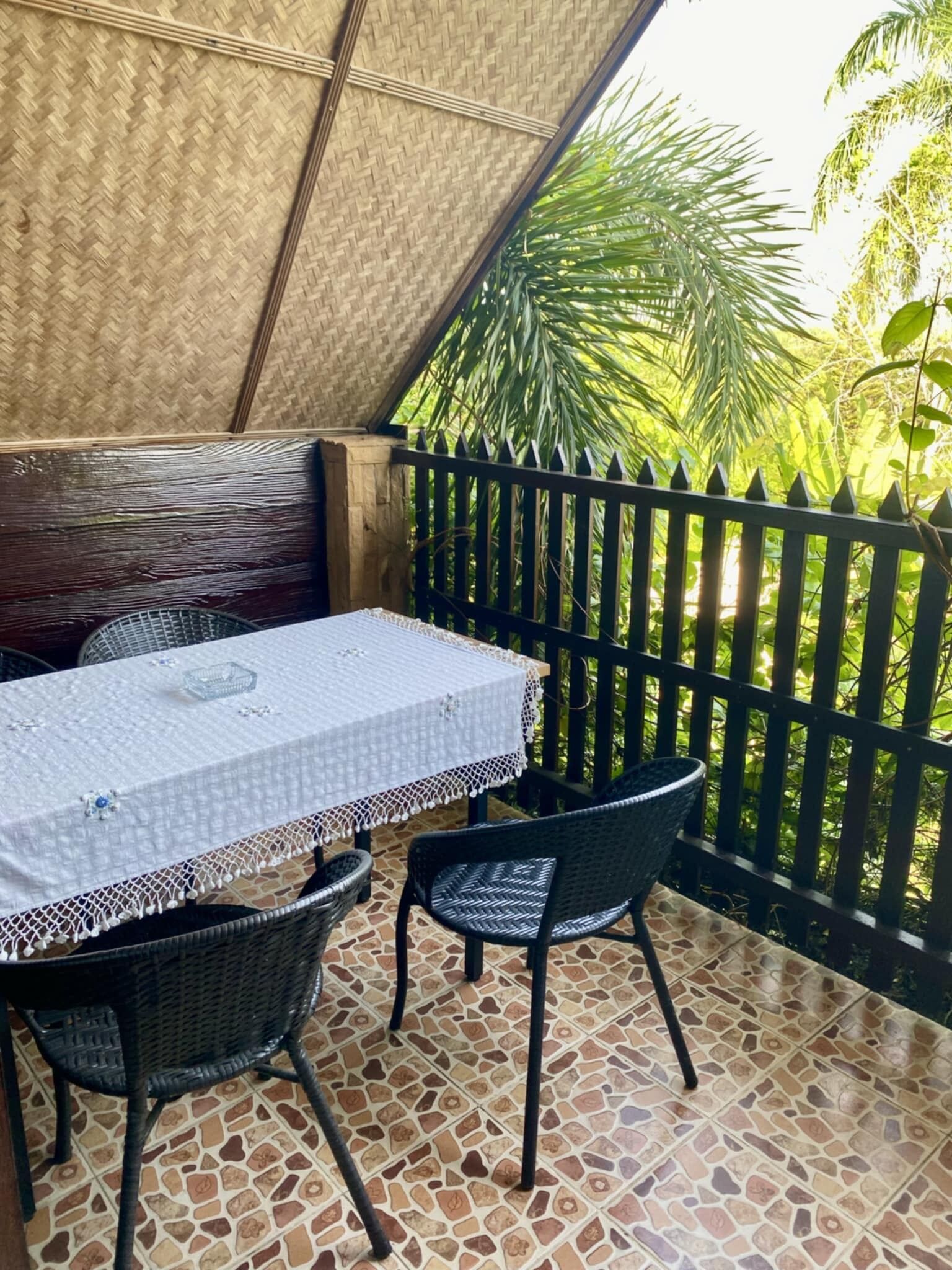 An outdoor patio with a table and chairs, a woven ceiling, and a view of greenery.