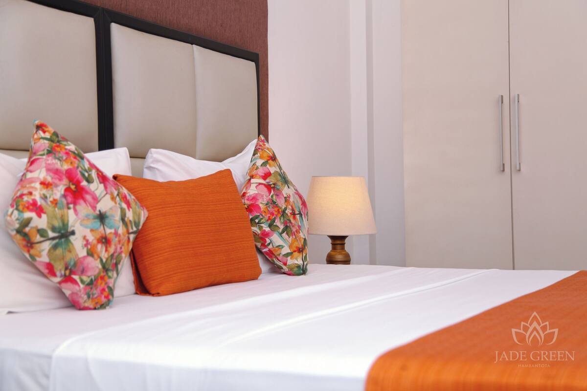 Un lit bien fait, avec une literie blanche et des coussins aux motifs floraux et de couleur orange, une lampe de chevet et une armoire au design simple.