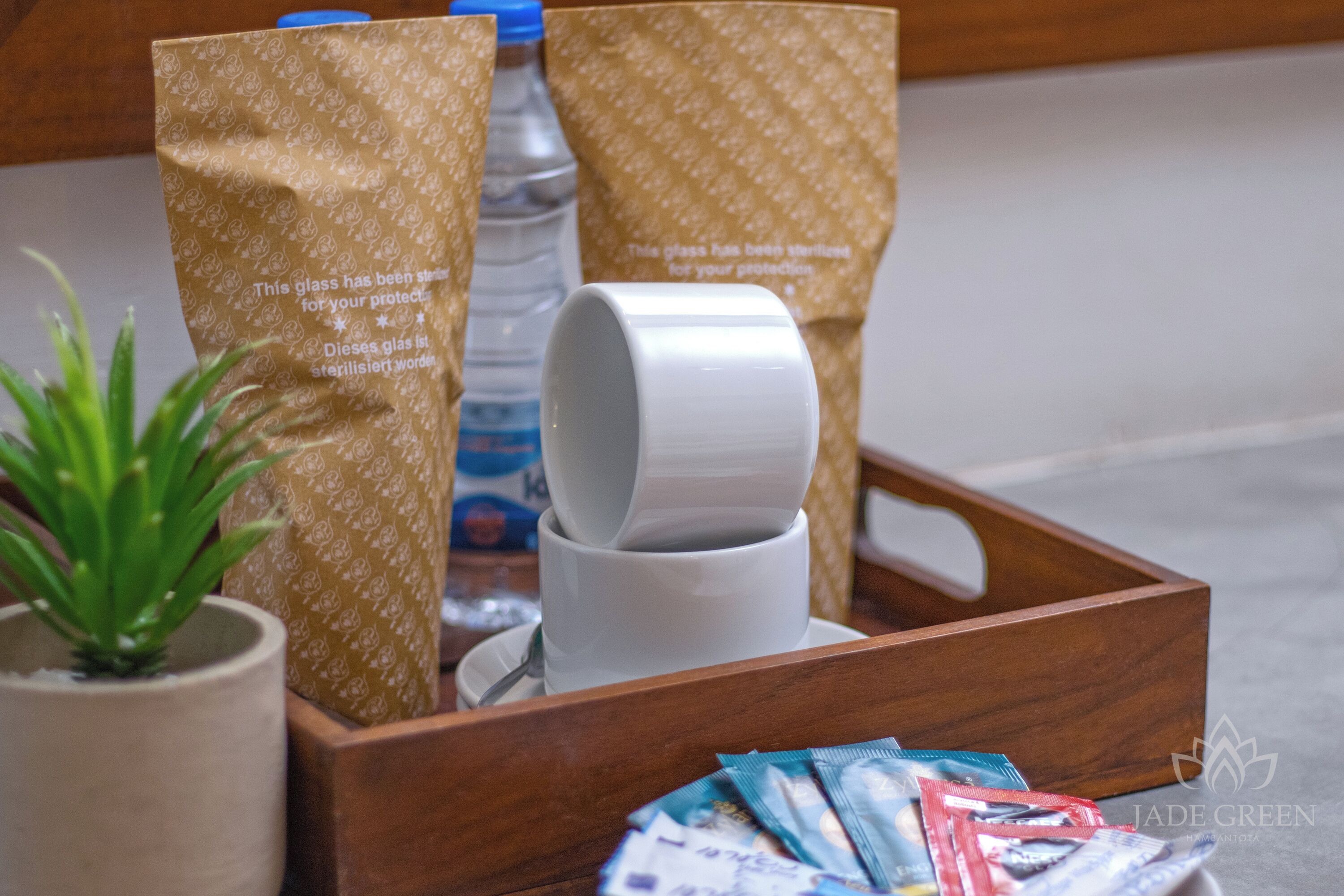 Ein Holztablett mit einer weißen Tasse, zwei gemusterten Papiertüten, einer Wasserflasche und einem kleinen Topfpflanzen.