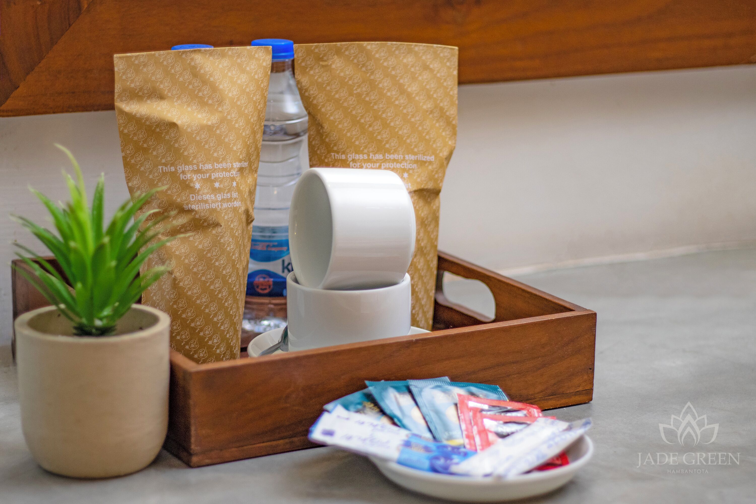 A wooden tray with a potted plant, a bottle of water, a roll of toilet paper, and a plate with packets.