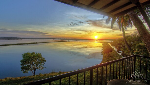 Vista al atardecer desde un balcón cubierto, con vista a un lago tranquilo y una orilla lejana con palmeras.