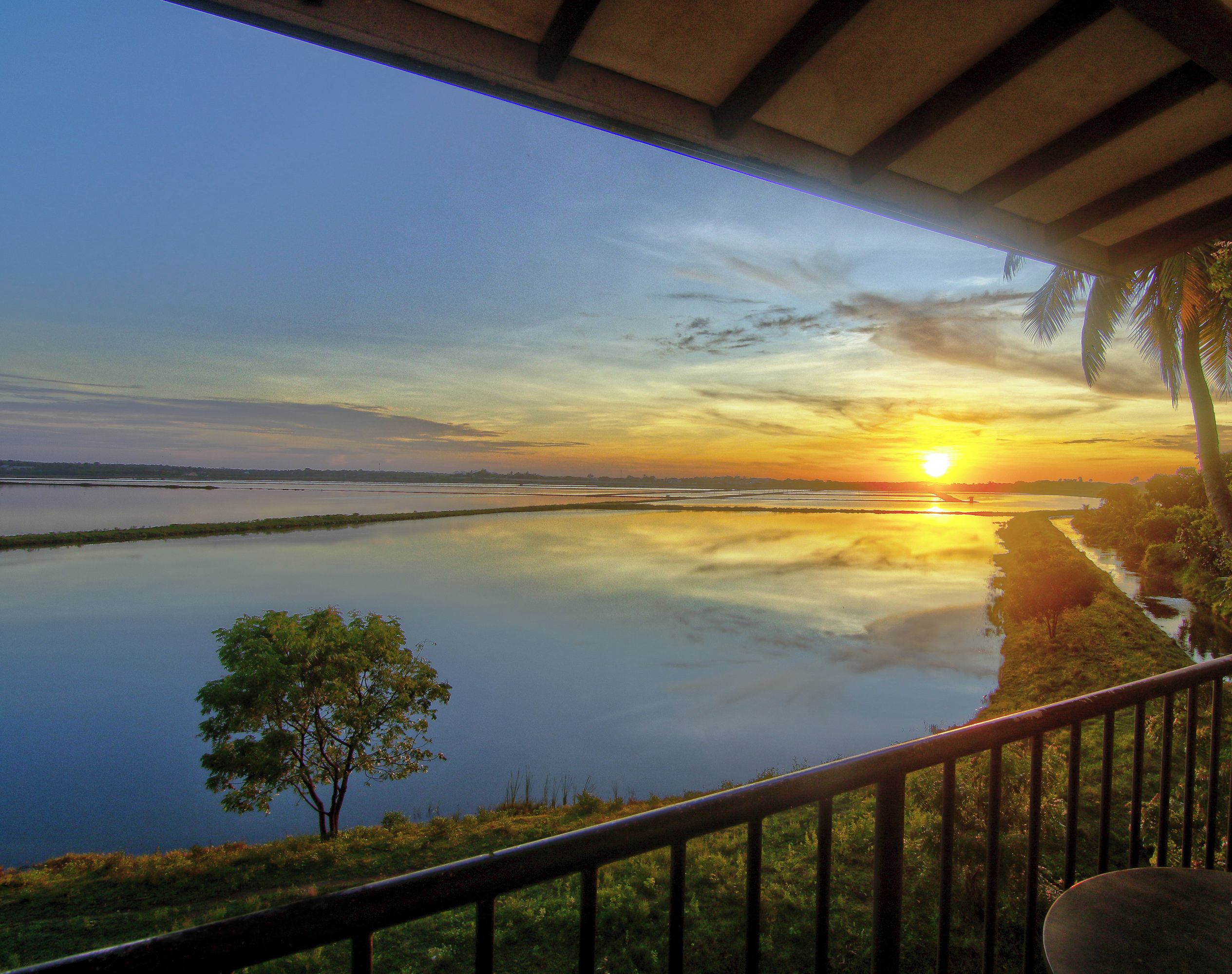 Vista al atardecer desde un balcón cubierto, con vista a un lago tranquilo y una orilla lejana con palmeras.
