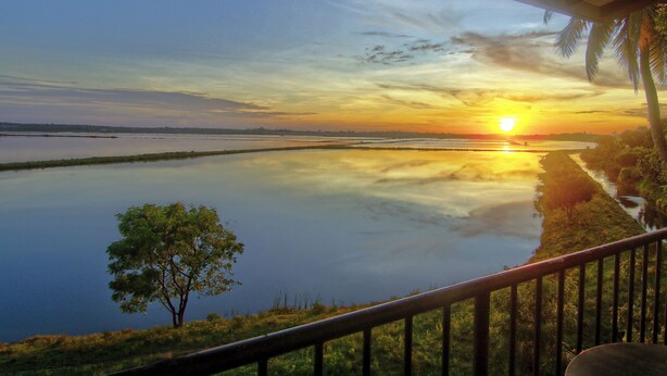 Une vue sur le coucher de soleil depuis un balcon abrité, donnant sur un lac paisible, avec une rive lointaine et des palmiers.