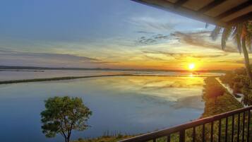 Vista do pôr do sol a partir de uma varanda coberta, com vista para um lago calmo, costa distante e palmeiras.