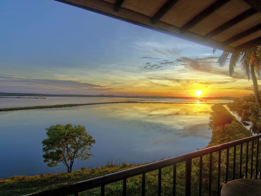 A sunset view from a covered balcony overlooking a calm lake with a distant shoreline and palm trees.