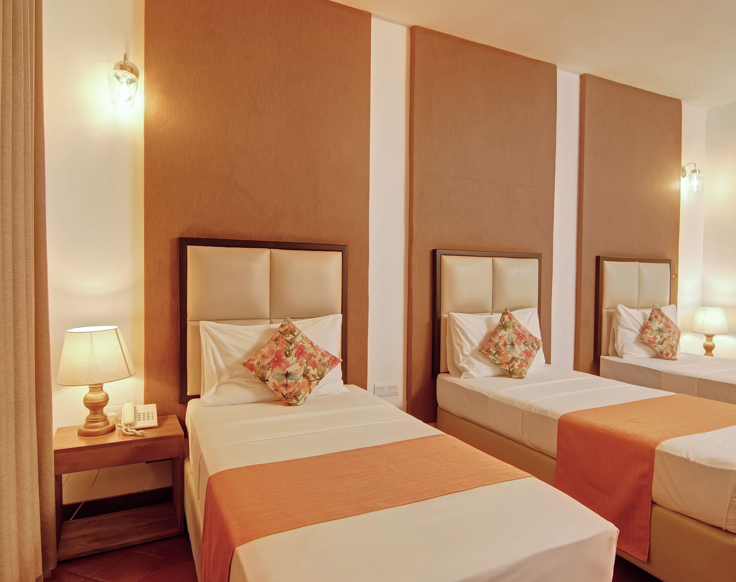 Une chambre d’hôtel avec trois lits, chacun doté d’une tête de lit et d’un oreiller. On y trouve une table de chevet avec une lampe et un téléphone.