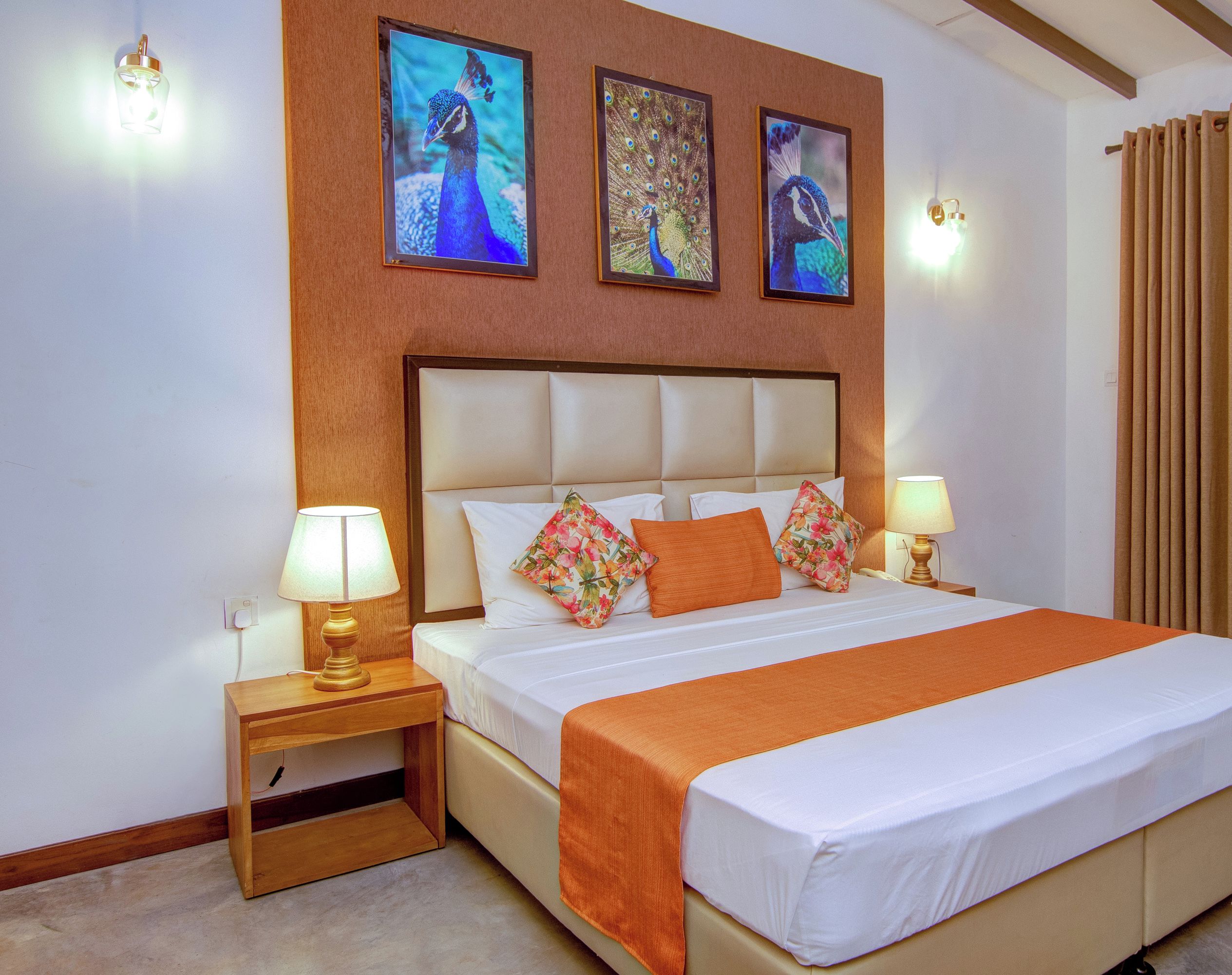 A neatly made bed with a white and orange bedspread, flanked by two nightstands with lamps, and three framed peacock images on the wall above.