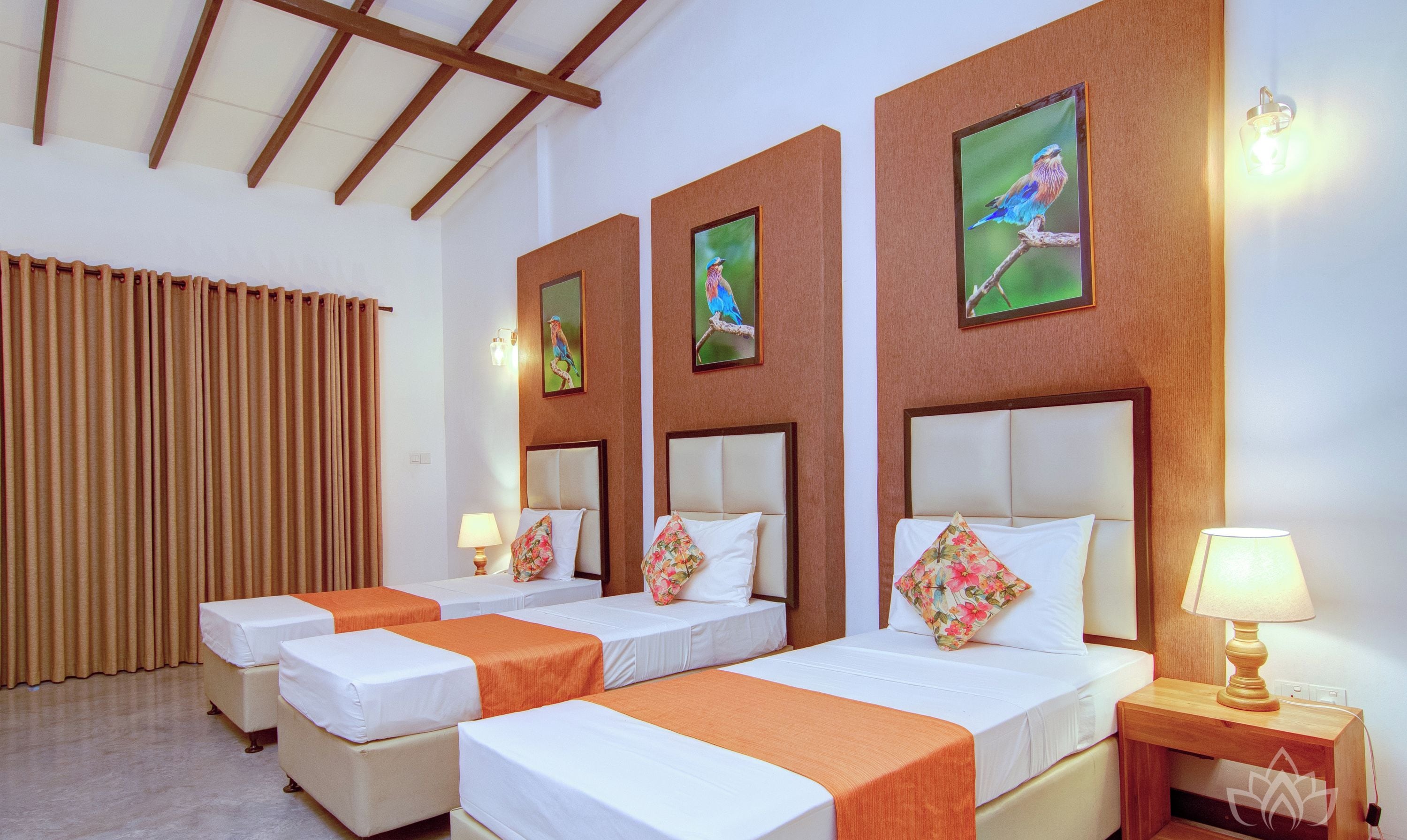 A hotel room with three single beds, each with an orange bedspread and white bedding. There are two wall-mounted paintings of birds, a wooden headboard, and a nightstand with a lamp.