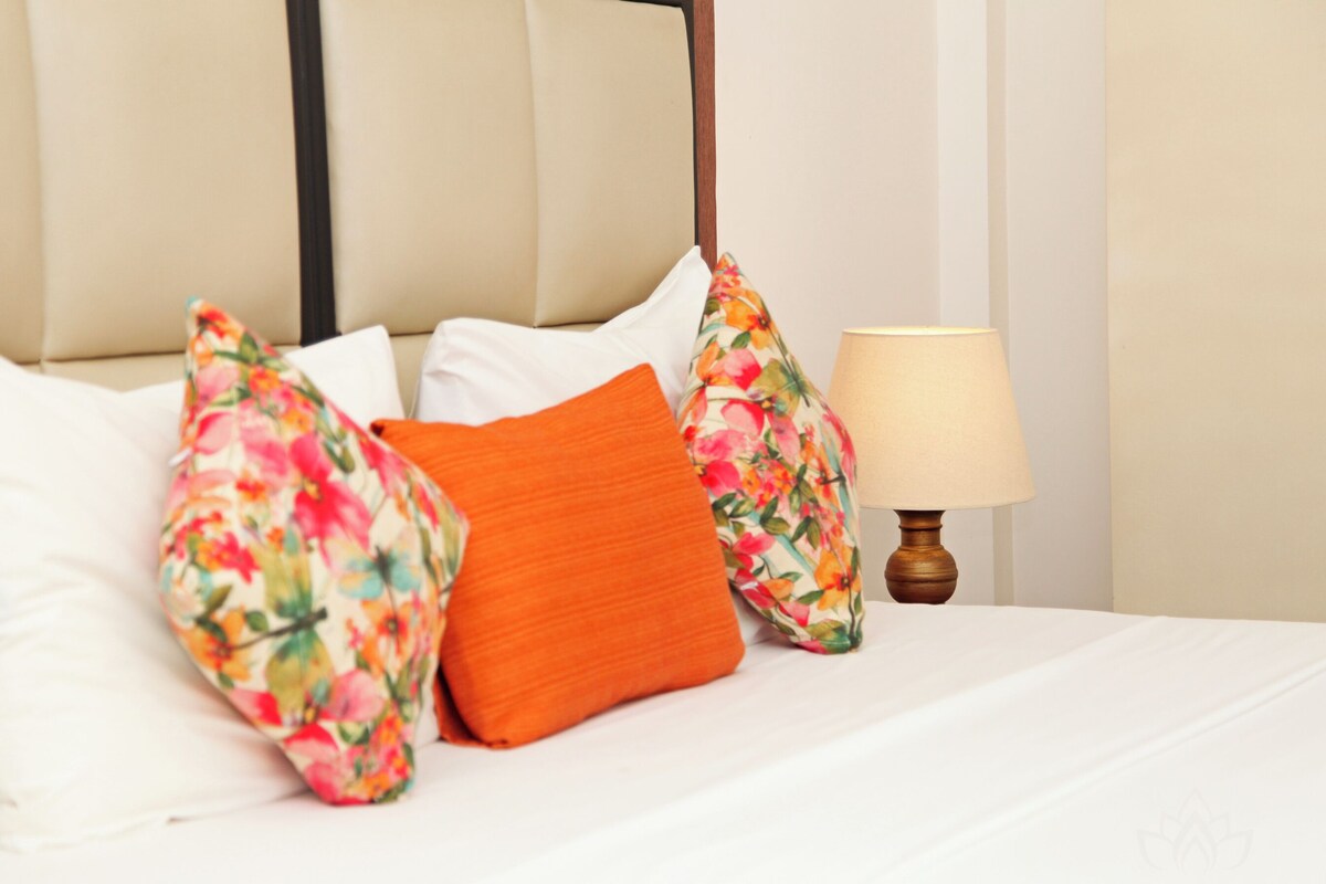 Una cama bien tendida con almohadas blancas y anaranjadas, una almohada estampada con motivos florales y una lámpara de noche.