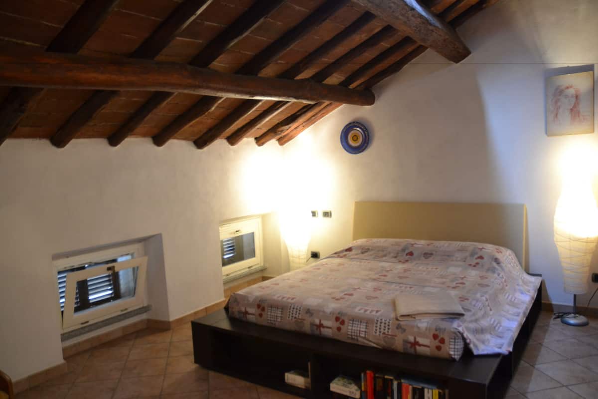 Un dormitorio con cama, ventana, televisor montado en la pared, lámpara y techo con vigas a la vista.