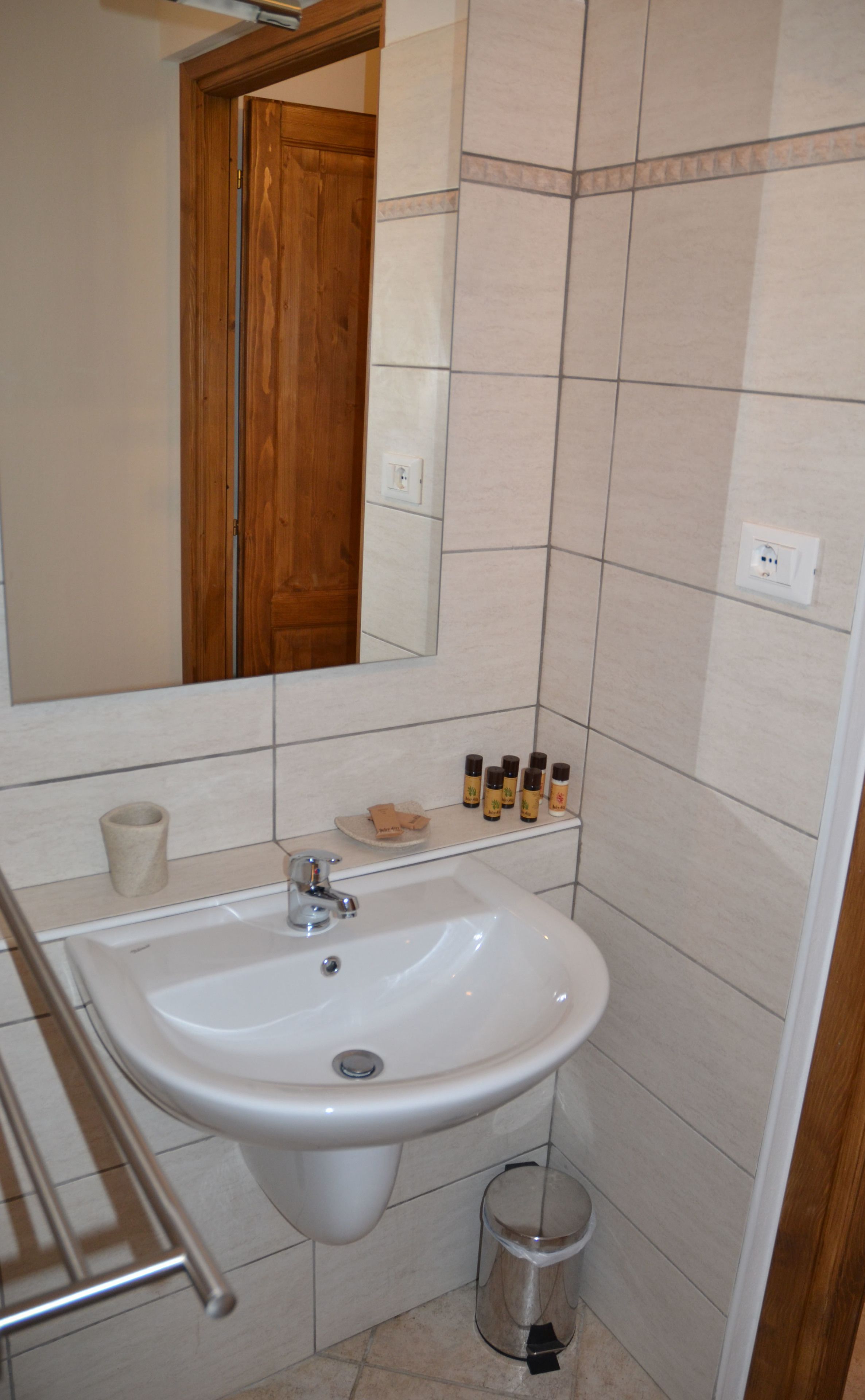 Un baño con un lavamanos blanco, un espejo y una puerta de madera. Hay artículos de aseo en el lavamanos y un cubo de basura a su lado.