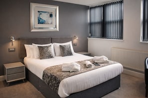 A neatly made bed with a patterned bedspread, a bedside table with a lamp, and a window with blinds. - The Royal Seabank Hotel (Blackpool)