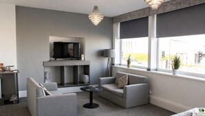 A modern living room with grey sofas, a flat-screen TV, a fireplace, and large windows with a view of the outside.