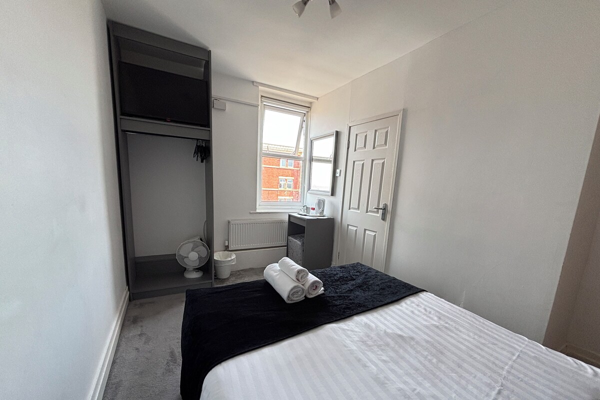 A bedroom with a bed, a window, a radiator, a fan, and a door.