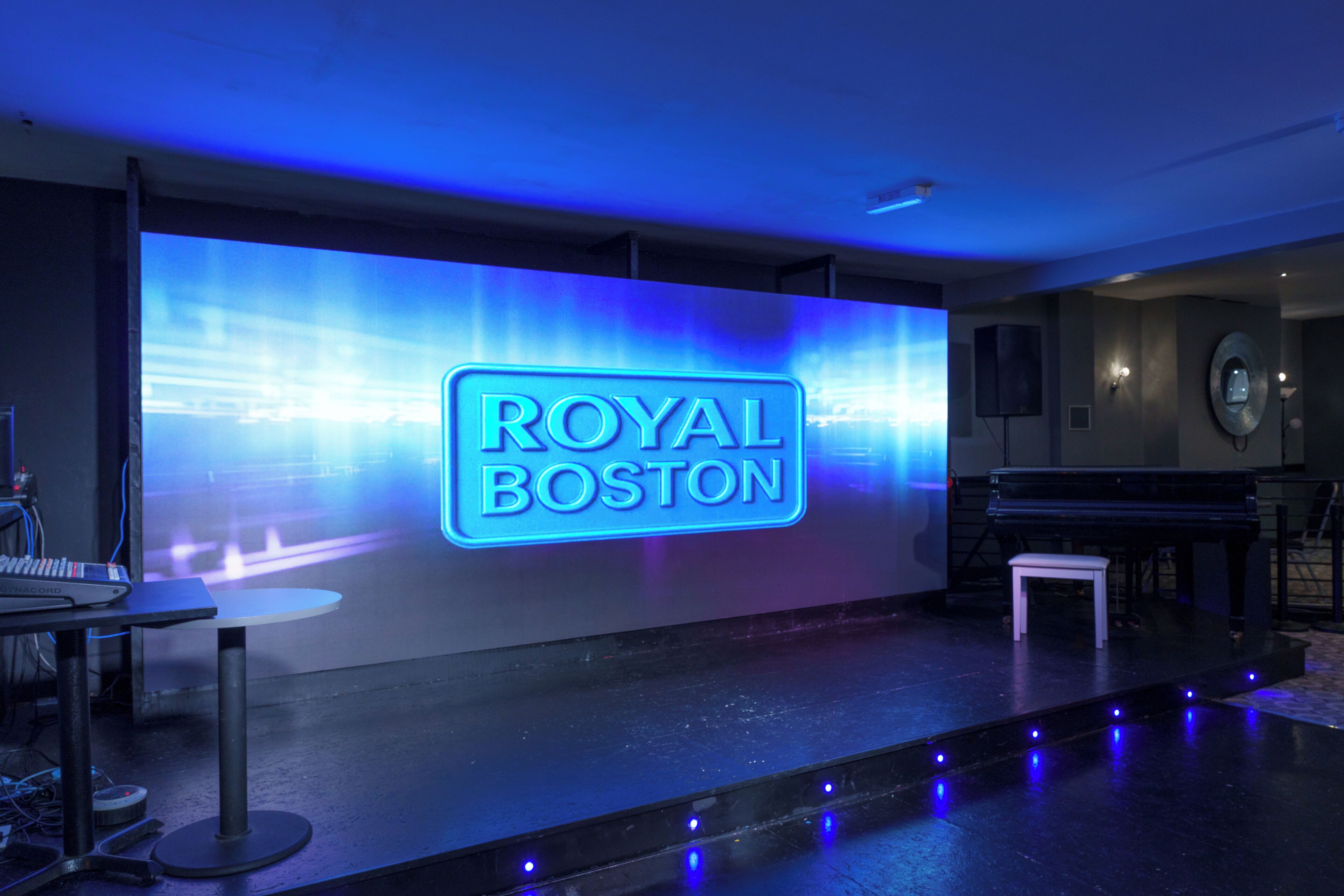 A stage with a neon sign reading "ROYAL BOSTON" and a piano on the right.